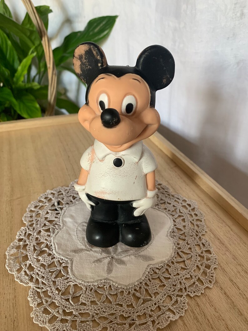 May include: Vintage Mickey Mouse figurine. The figure is wearing a white shirt with a black button, black shorts, and white gloves. The ears are black, and the face is tan. The figurine is standing on a gray doily.
