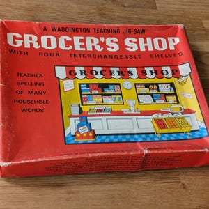 May include: A vintage Grocer's Shop jigsaw puzzle box. The red box features the title "GROCER'S SHOP" in large white letters, with an illustration of a grocery store. The box also states that it teaches spelling of household words.