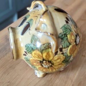 Può includere: Un salvadanaio in ceramica con un design floreale. Il salvadanaio è dipinto in tonalità di marrone, giallo e verde. I fiori sono dipinti in uno stile realistico.