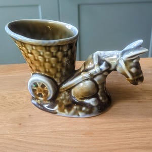 Peut inclure: Un âne en céramique brun et blanc tirant un chariot, avec une finition émaillée brun et blanc. Le chariot a un panier brun et blanc et une seule roue.