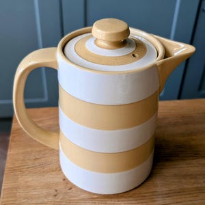 Puede incluir: Jarra de cerámica a rayas blancas y amarillo claro. Tiene un asa curva y un pico vertedor. La tapa tiene un pequeño pomo en la parte superior. Un artículo de cocina.