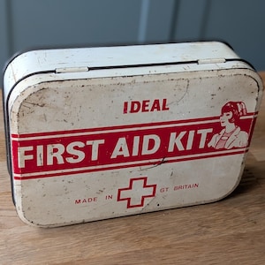 May include: A distressed white metal first aid kit with red lettering. The text "IDEAL FIRST AID KIT" is displayed, along with a red cross symbol and an illustration of a woman. The kit is marked "MADE IN GT BRITAIN".