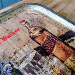 May include: Vintage metal serving tray with a distressed finish. The tray features a graphic of a Johnnie Walker whiskey bottle and glasses. The words "Johnnie Walker" are printed in red on the tray.