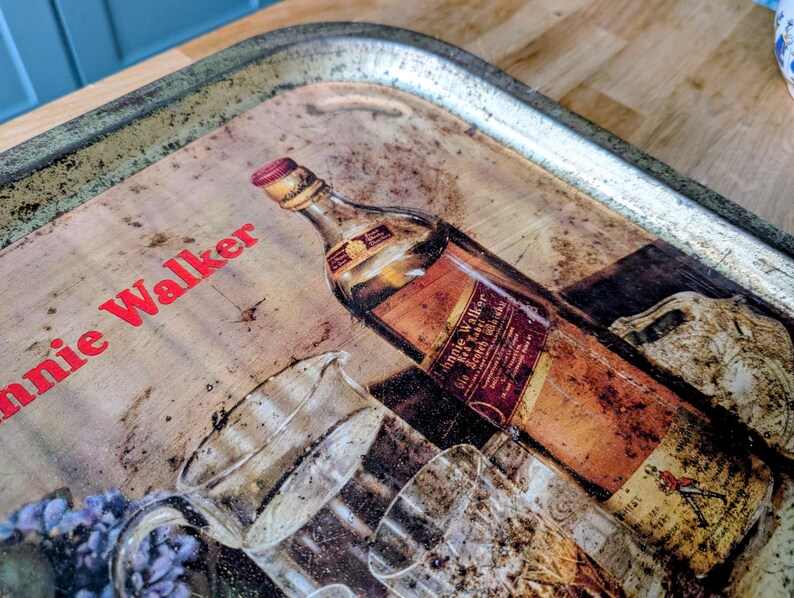 May include: Vintage metal serving tray with a distressed finish. The tray features a graphic of a Johnnie Walker whiskey bottle and glasses. The words "Johnnie Walker" are printed in red on the tray.