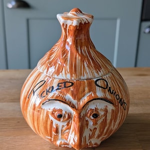 May include: A ceramic onion-shaped jar with a lid, painted in shades of orange and white. The jar has a face design and the words "Pickled Onions" are written on it. The jar is sitting on a wooden surface.