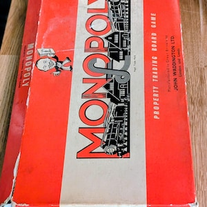 Op de afbeelding: Een vintage Monopoly bordspeldoos, voornamelijk rood met een witte streep in het midden. Het woord "MONOPOLY" staat vetgedrukt. De doos bevat illustraties en tekst, die aangeven dat het een vastgoedhandel bordspel is.
