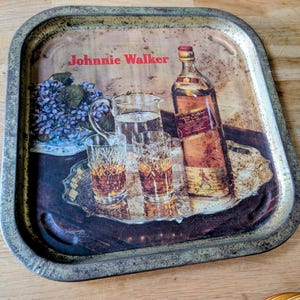 May include: Vintage rectangular metal tray with a Johnnie Walker whiskey advertisement. The tray features a bottle of whiskey, glasses with amber liquid, and a floral arrangement. The tray has a distressed, aged appearance.