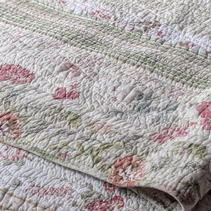 May include: A quilted bedspread featuring a floral pattern with pink and red blossoms, green foliage, and white background sections. The fabric has a textured, stitched surface with horizontal striped borders in muted green and white tones.