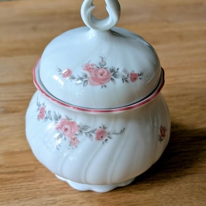 May include: White porcelain sugar bowl with a lid, decorated with pink roses and a pink rim. The lid has a loop handle. The bowl has a spiral design. A decorative piece for serving sugar or other condiments.