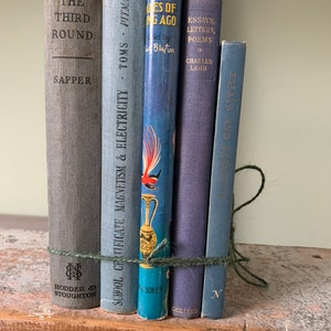 Könnte beinhalten: Ein Stapel von sechs blauen und grauen gebundenen Büchern mit einem grünen Bindfaden, der um sie herumgebunden ist. Die Bücher sind betitelt mit "The Third Round", "School Certificate Magnetism & Electricity", "Tales of Long Ago", "Essays, Letters, Poems" und "Essays and Stories".
