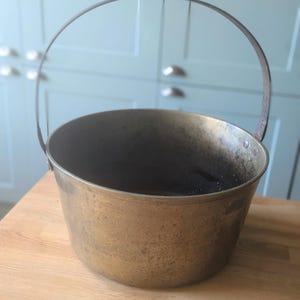 Può includere: Una pentola in ottone vintage con un manico in metallo. La pentola ha un aspetto usurato e invecchiato ed è leggermente opaca.