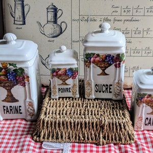 Può includere: Quattro barattoli in ceramica bianca con coperchi, ognuno decorato con una ciotola di frutta e dettagli floreali. I barattoli sono etichettati con "Farine", "Poivre", "Sucre" e "Cafe".
