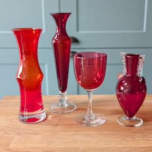 May include: Four red glass vases of different shapes and sizes. The vases are all made of clear glass and have a deep red colour. The vases are arranged on a wooden surface.