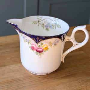 May include: A white porcelain creamer with a navy blue rim and gold detailing. The creamer has a floral pattern with pink, yellow, and purple flowers. The handle is curved and the spout is pointed.