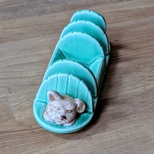 May include: A turquoise ceramic bread knife holder with a dog's head on the front. The holder has four slots for bread knives.