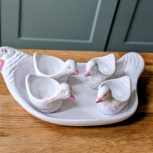 Può includere: Un piatto da portata in ceramica bianca con quattro portauova a forma di anatra. Il piatto ha una testa d'anatra a un'estremità e una coda all'altra. I portauova e le teste d'anatra hanno becchi rosa.