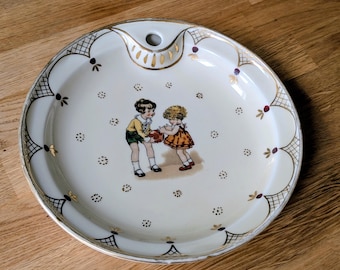 May include: White ceramic plate with a decorative gold rim and a handle. The plate features a painted image of two children playing with a ball. Gold and purple accents are scattered across the plate's surface.