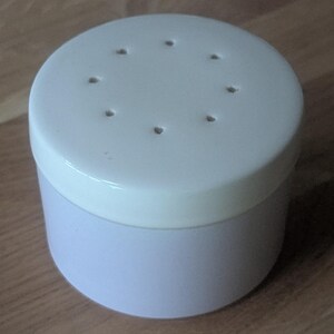 May include: A white ceramic container with a lid. The lid has a circle of small holes around the center. The container is a light purple color.