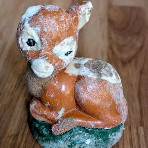 May include: A small, ceramic figurine of a fawn sitting on a green base. The fawn is brown with white markings and has black eyes. The figurine is covered in a layer of white dust.
