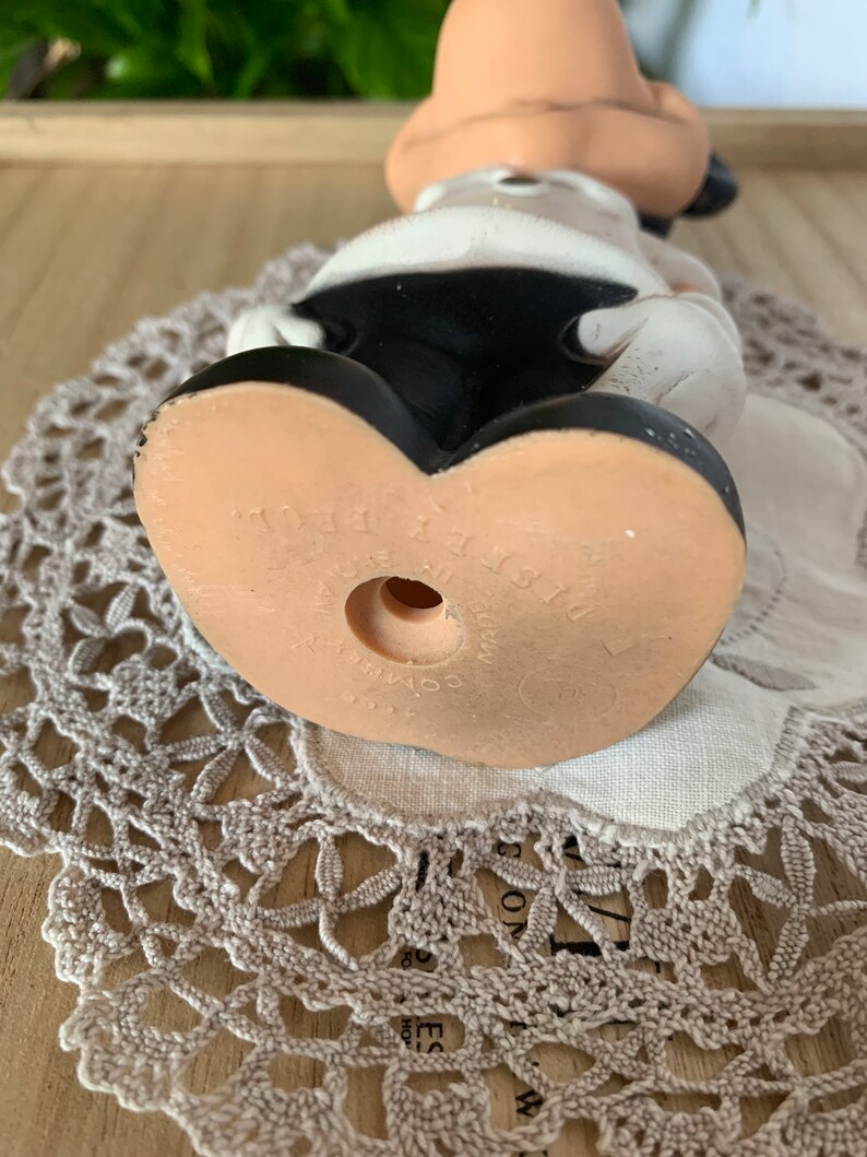 May include: A vintage Mickey Mouse figurine, viewed from the bottom up, showcasing the character's iconic black shorts and large, tan feet. The figurine is made of a light-colored material, with the words "Disney Prod." and "Made in Japan" visible on the sole.