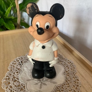 May include: A vintage Mickey Mouse figurine, featuring black ears, a black nose, and a white shirt with black pants. The figurine is standing on a lace doily, with a wooden tray and greenery in the background.