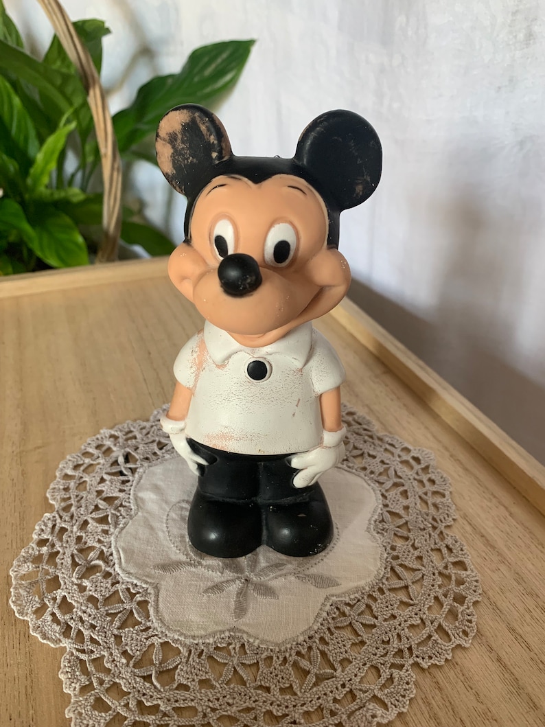 May include: A vintage Mickey Mouse figurine, featuring black ears, a black nose, and a white shirt with black pants. The figurine is standing on a lace doily, with a wooden tray and greenery in the background.