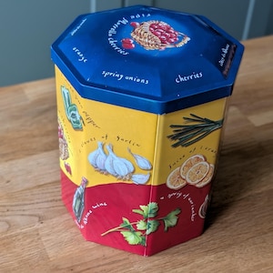 May include: An octagonal tin with a blue lid and yellow and red body. The lid features text and an illustration of cherries. The body has illustrations of garlic, herbs, and other ingredients. Text includes "cherries", "spring onions", and "cloves of garlic".