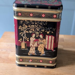 Puede incluir: Lata rectangular negra con un diseño de inspiración asiática. La tapa roja y dorada presenta un motivo de pájaro. Los lados muestran una escena con figuras, árboles y una planta en maceta, todo en dorado sobre fondo negro.