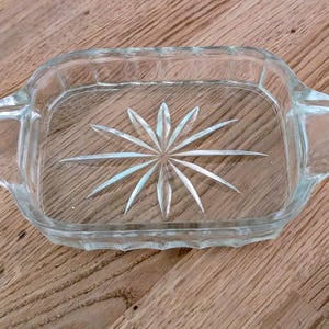 May include: Clear glass rectangular dish with two handles. The dish features a starburst design in the centre. The dish is made of clear glass and is sitting on a wooden surface.