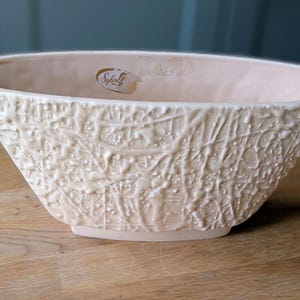 May include: Cream-coloured, oval ceramic bowl with a textured, raised pattern. The bowl has a wide opening and a flat base. A small, faded sticker is visible near the rim. Likely for decorative or serving purposes.