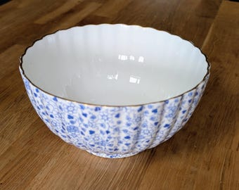 Pode incluir: Uma tigela de cerâmica branca com um padrão floral azul e uma borda dourada. A tigela tem uma borda ondulada e está sobre uma superfície de madeira. A tigela tem cerca de 20 cm de diâmetro.