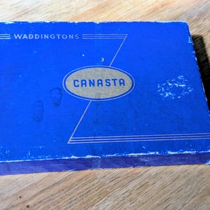 Könnte beinhalten: Eine blaue Vintage-Kartenspielbox von Waddington's Canasta. Die Box hat goldfarbene Schriftzüge und Designelemente, mit dem Wort "CANASTA" in einem Oval. Die rechteckige Box weist Gebrauchsspuren auf.
