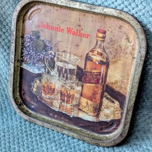 May include: Vintage metal serving tray with a distressed finish. The tray features a bottle of Johnnie Walker Scotch Whisky, glasses, and a pitcher. The text "Johnnie Walker" is printed in red.