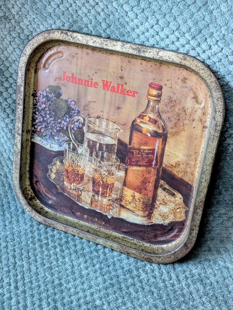 May include: Vintage metal serving tray with a distressed finish. The tray features a bottle of Johnnie Walker Scotch Whisky, glasses, and a pitcher. The text "Johnnie Walker" is printed in red.