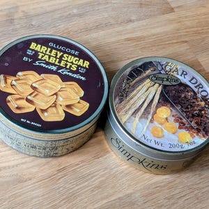 Puede incluir: Dos latas redondas de metal de caramelos. La lata de la izquierda es dorada con una etiqueta roja que dice "Glucose Barley Sugar Tablets by Smith Kendon" y presenta una imagen de cubos de azúcar dorados. La lata de la derecha es dorada con una etiqueta roja que dice "Barley Sugar Drops Simpkins" y presenta una imagen de cebada y cristales de azúcar marrón.