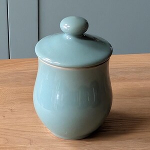 May include: A light blue ceramic jar with a lid. The jar has a rounded shape and a smooth, glossy finish.