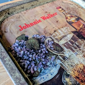 May include: Vintage metal serving tray with a distressed finish. The tray features a print of a Johnnie Walker bottle, glasses, and a floral arrangement. The text "Johnnie Walker" is printed in red across the top.