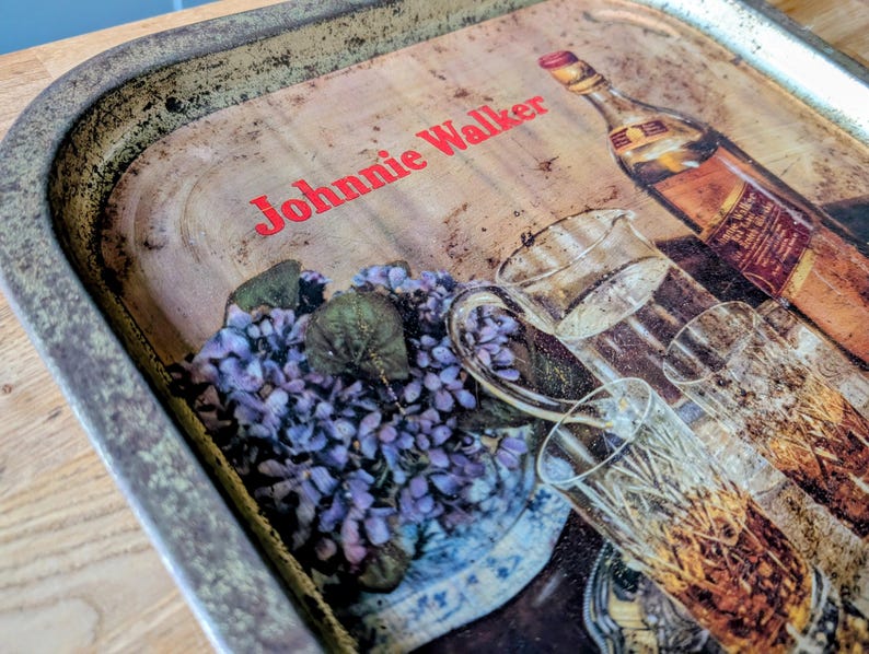 May include: Vintage metal serving tray with a distressed finish. The tray features a print of a Johnnie Walker bottle, glasses, and a floral arrangement. The text "Johnnie Walker" is printed in red across the top.
