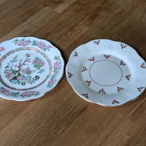 Pode incluir: Dois pratos decorativos de porcelana. Um prato apresenta um design floral com flores rosa e azuis, uma árvore e uma borda dourada. O outro é branco com uma borda dourada e detalhes florais vermelhos. Ambos os pratos têm bordas onduladas.