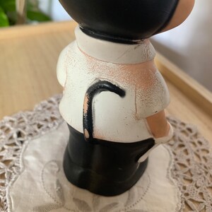 May include: A vintage figurine of a person in a black suit and hat, with a white shirt and a black cane. The figurine is placed on a white doily with a floral pattern. The figure has a black head and a white shirt.