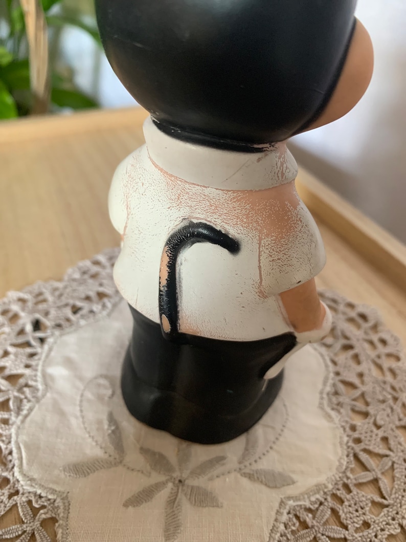 May include: A vintage figurine of a person in a black suit and hat, with a white shirt and a black cane. The figurine is placed on a white doily with a floral pattern. The figure has a black head and a white shirt.