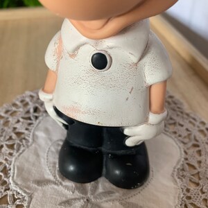May include: A figurine of a cartoon character wearing a white shirt with a black button, black pants, and white gloves. The figurine has a distressed, vintage appearance. The figure is standing on a white, embroidered doily.