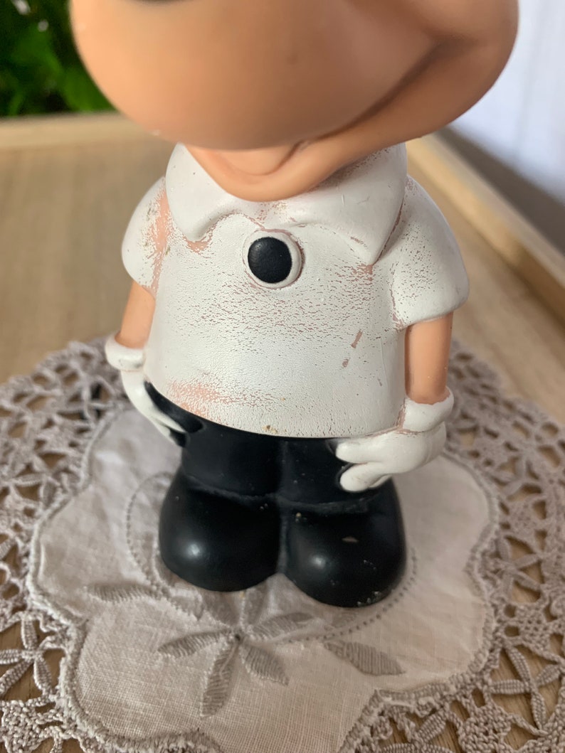 May include: A figurine of a cartoon character wearing a white shirt with a black button, black pants, and white gloves. The figurine has a distressed, vintage appearance. The figure is standing on a white, embroidered doily.