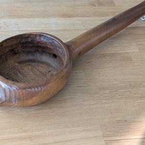 May include: A hand-carved wooden ladle with a deep bowl and long handle. The ladle is made of a dark brown wood with visible grain and a smooth finish. A small metal hook is attached to the end of the handle.