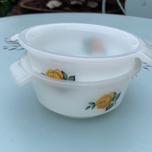 May include: Two white ceramic bowls with yellow rose designs. The bowls are stacked on top of each other. The bowl on top has a lid.