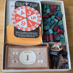May include: A vintage Monopoly board game box containing a red and white circular spinner with numbers, a stack of orange cards, a compartment filled with small teal and maroon wooden houses, and a stack of brown paper money with a rocking horse token.
