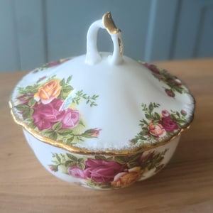 以下が含まれることがあります： 金色の縁取りと蓋が付いた白い磁器製の砂糖入れ。赤い、ピンクの、そして黄色のバラの花柄で飾られています。