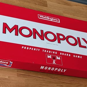 Puede incluir: Una caja rectangular del juego de mesa Monopoly con un diseño en rojo y blanco. La caja presenta el logotipo de Waddingtons, el título Monopoly en letras rojas grandes y texto que indica que es un juego de mesa para 2 a 6 jugadores.