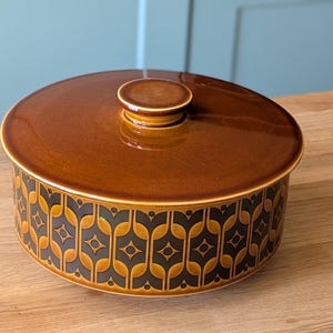 Puede incluir: Un plato de servir de cerámica marrón con tapa. El plato tiene un patrón geométrico en negro y marrón.