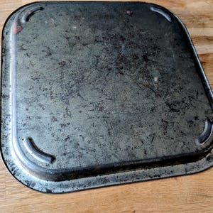 May include: A rectangular, vintage metal tray with rounded corners. The tray has a distressed, mottled gray finish with dark edges and visible scratches. The corners feature small, curved indentations.